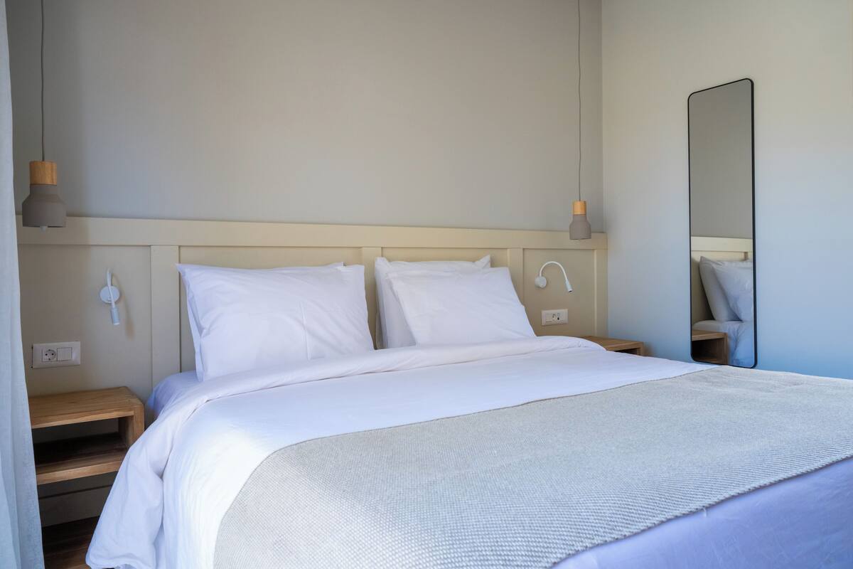 A neatly made bed with white bedding, a bedside table, a wall-mounted lamp, and a mirror.