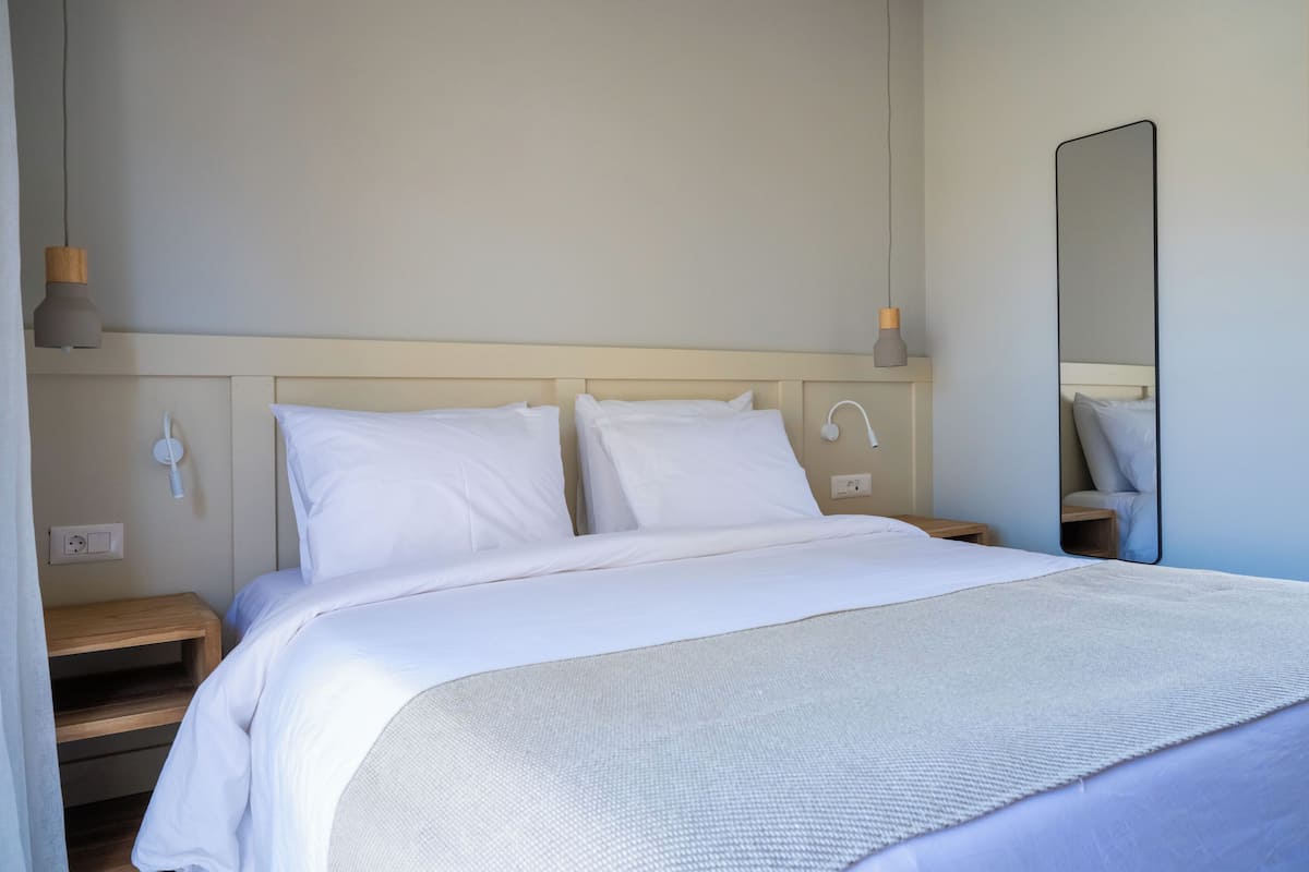 A neatly made bed with white bedding, a bedside table, a wall-mounted lamp, and a mirror.
