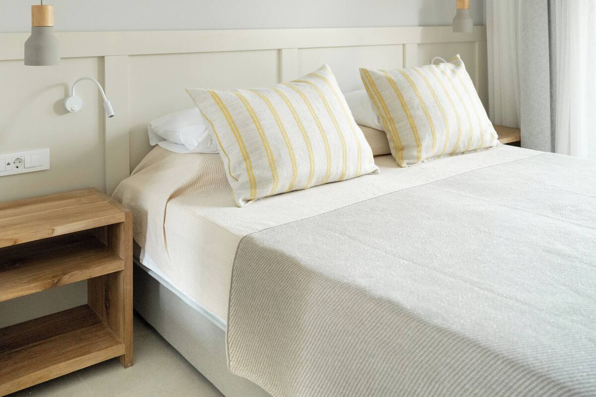 A neatly made bed with a wooden nightstand, a white wall, and a hanging light fixture.