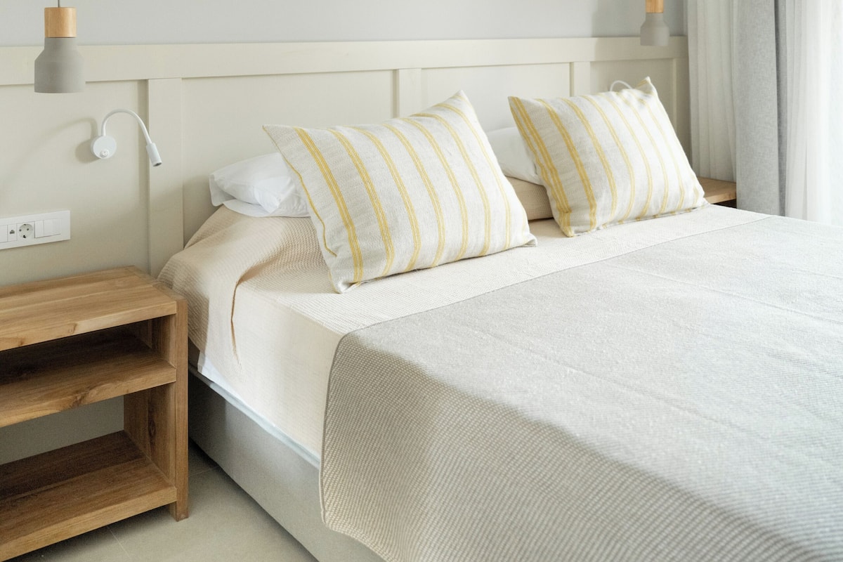 A neatly made bed with a wooden nightstand, a white wall, and a hanging light fixture.