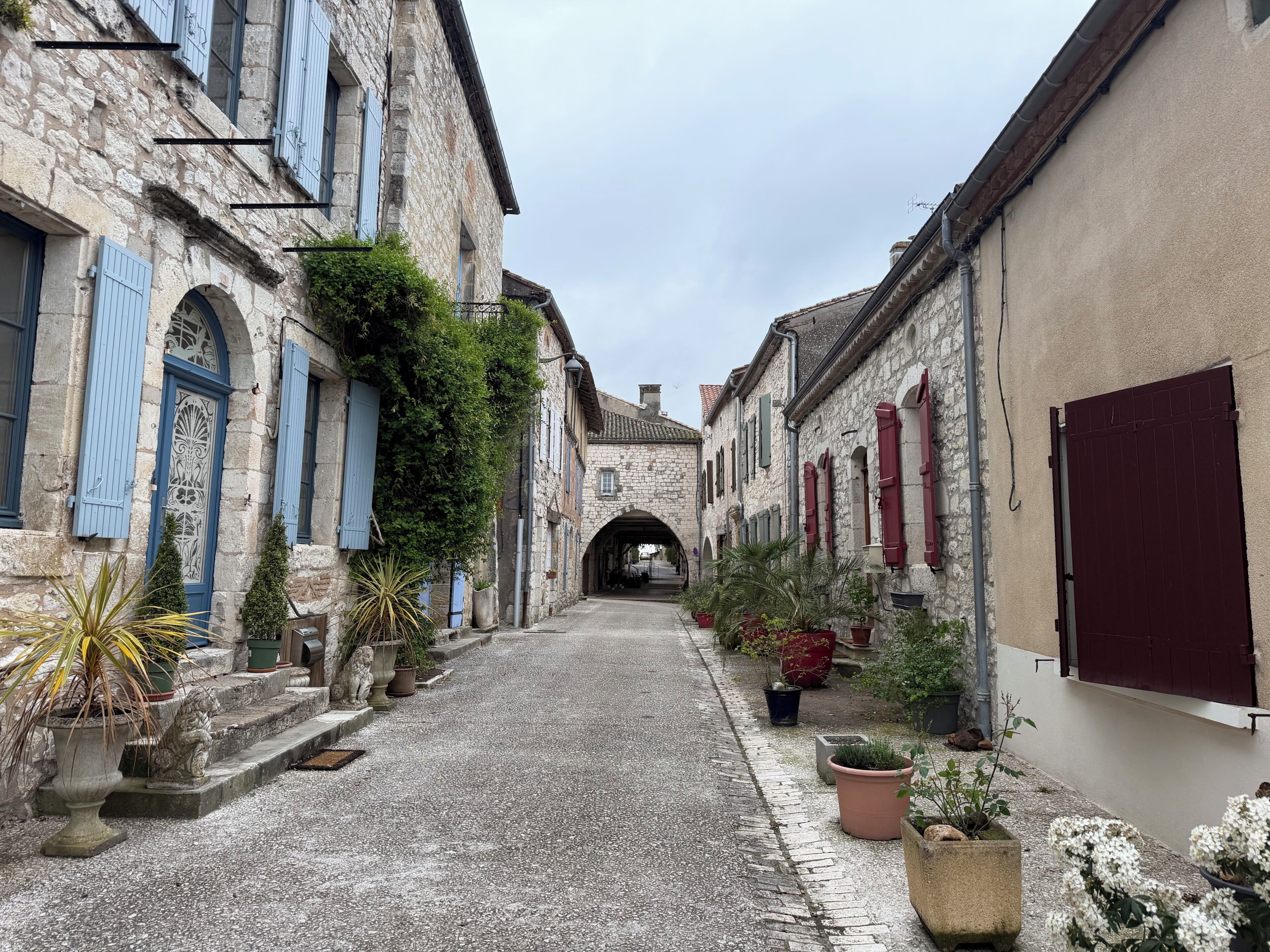 Une ruelle étroite pavée, bordée de bâtiments anciens en pierre, dotés d’arcades et de volets.