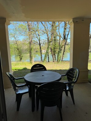 Eine überdachte Terrasse mit einem runden Tisch und vier Stühlen, mit Blick auf ein Gewässer und Bäume.