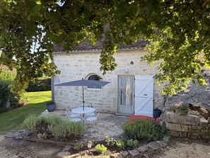 A stone building with a tiled roof, a white door, and a small garden with a table and chair under a canopy.