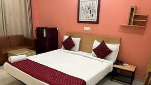 A hotel room with a bed, a nightstand, a wooden shelf, a red refrigerator, a brown sofa, and a framed picture on the wall. - Cloud Bay Hotels - Sector 69 (Gurugram)
