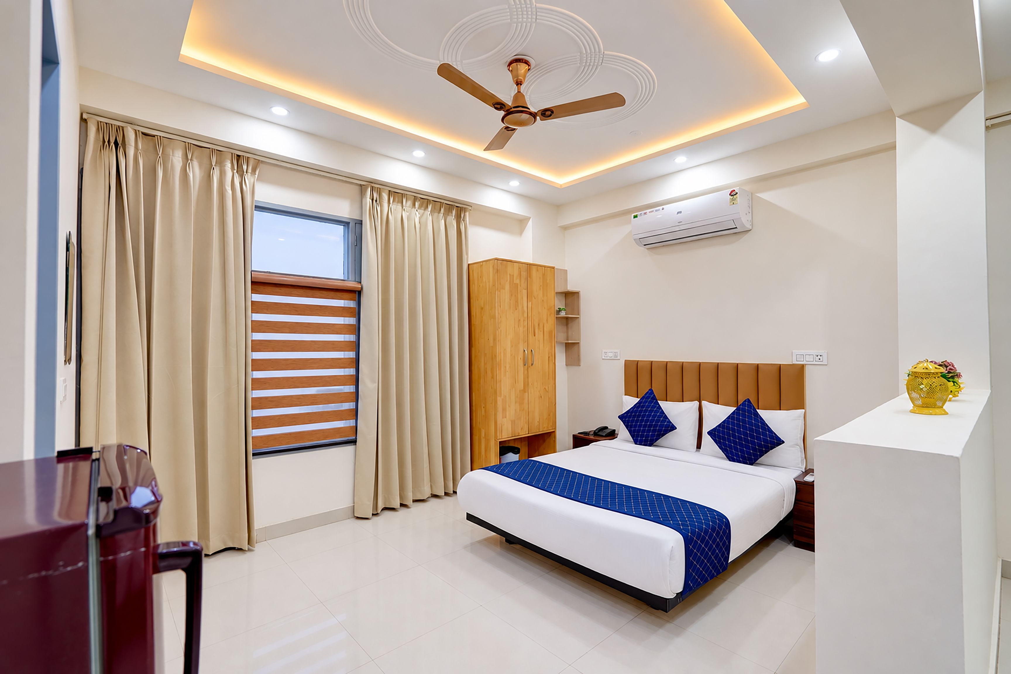 A hotel room with a bed, curtains, a ceiling fan, an air conditioning unit, and a bedside table.