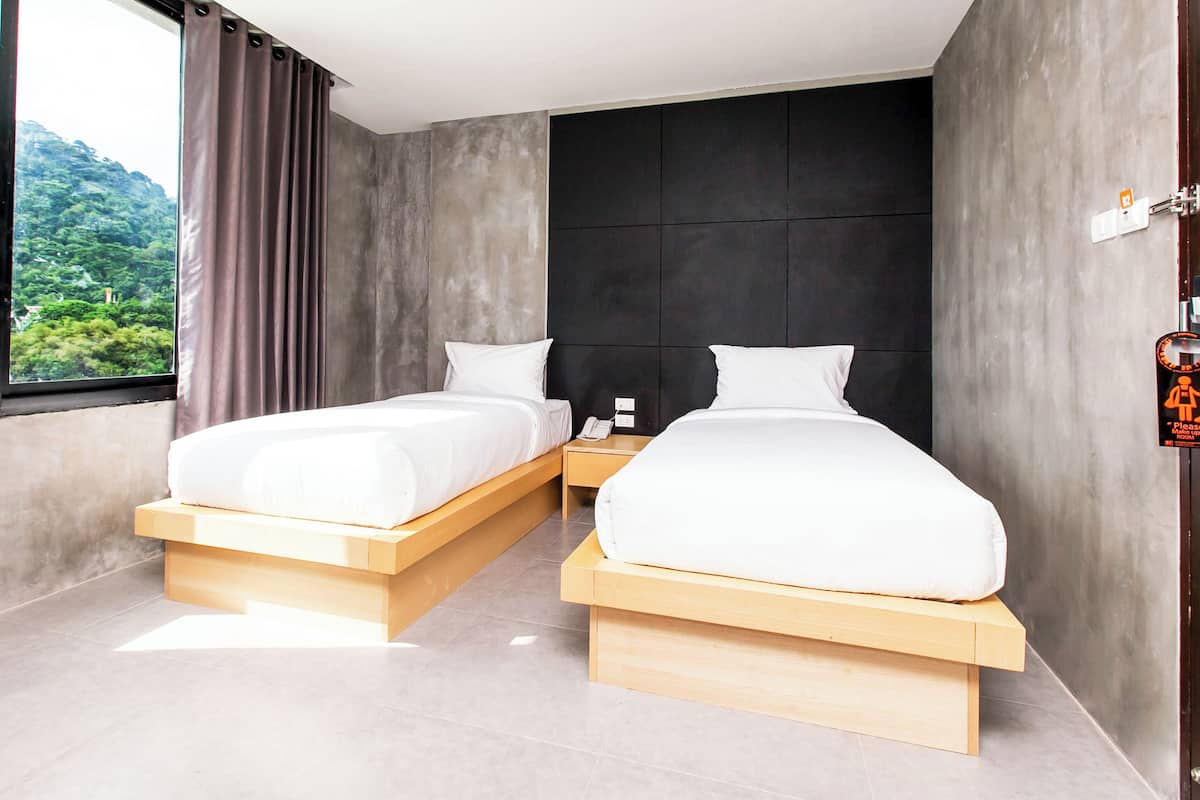 Una habitación de hotel moderna con dos camas, una plataforma de madera, una mesita de noche con un teléfono, vistas a zonas verdes y una puerta con un cartel.