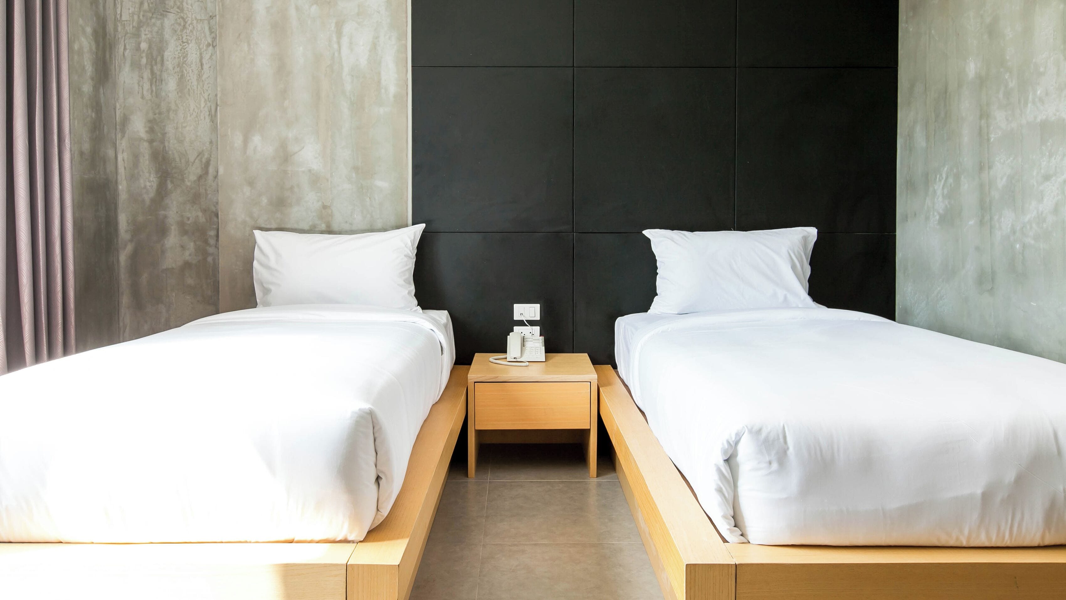 A hotel room with two single beds, a small wooden nightstand with a phone, and a concrete wall with a black panel.