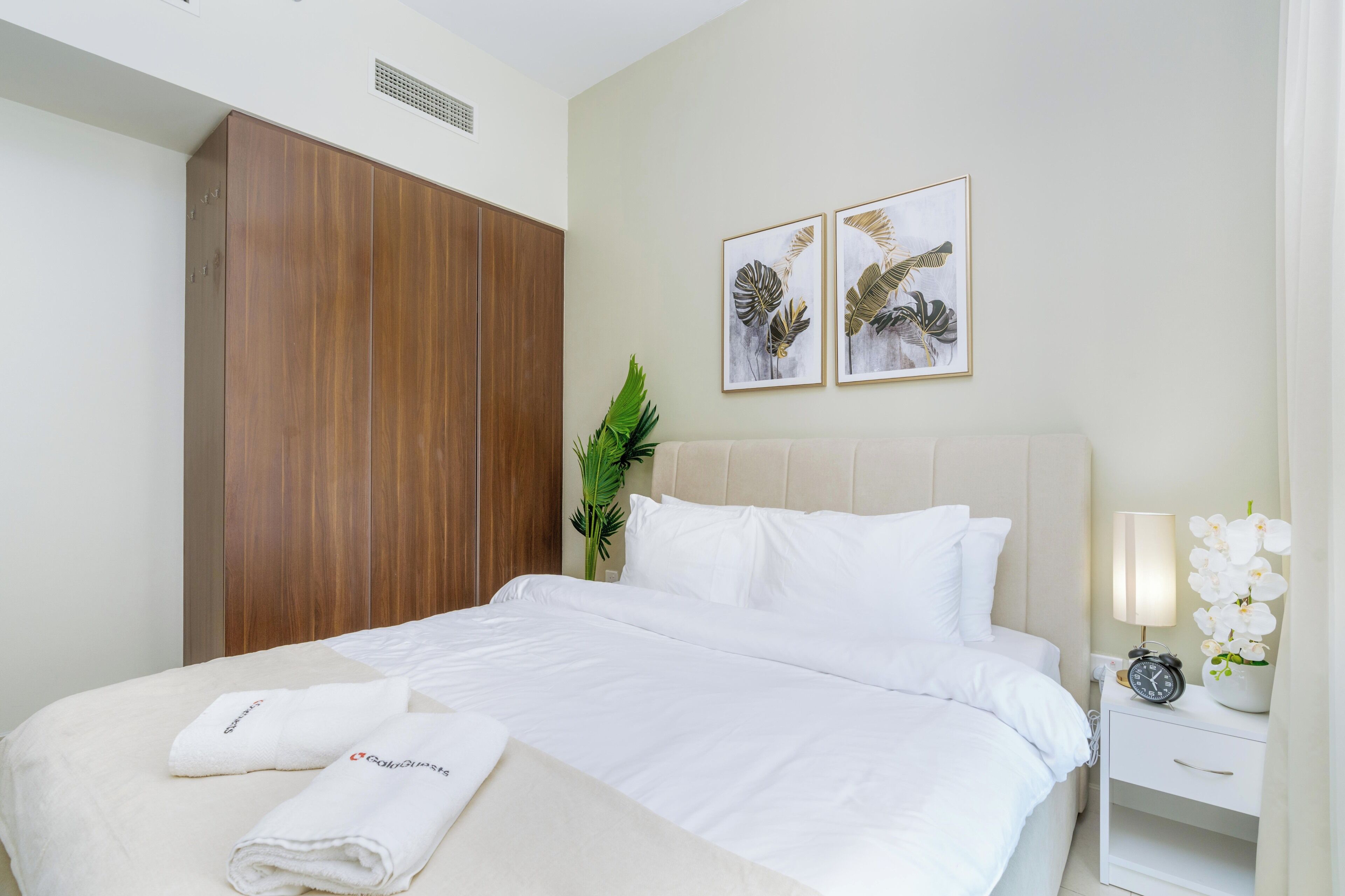 A neatly made bed with white linens and a folded towel, a bedside table with a lamp and a clock, and a large wardrobe.