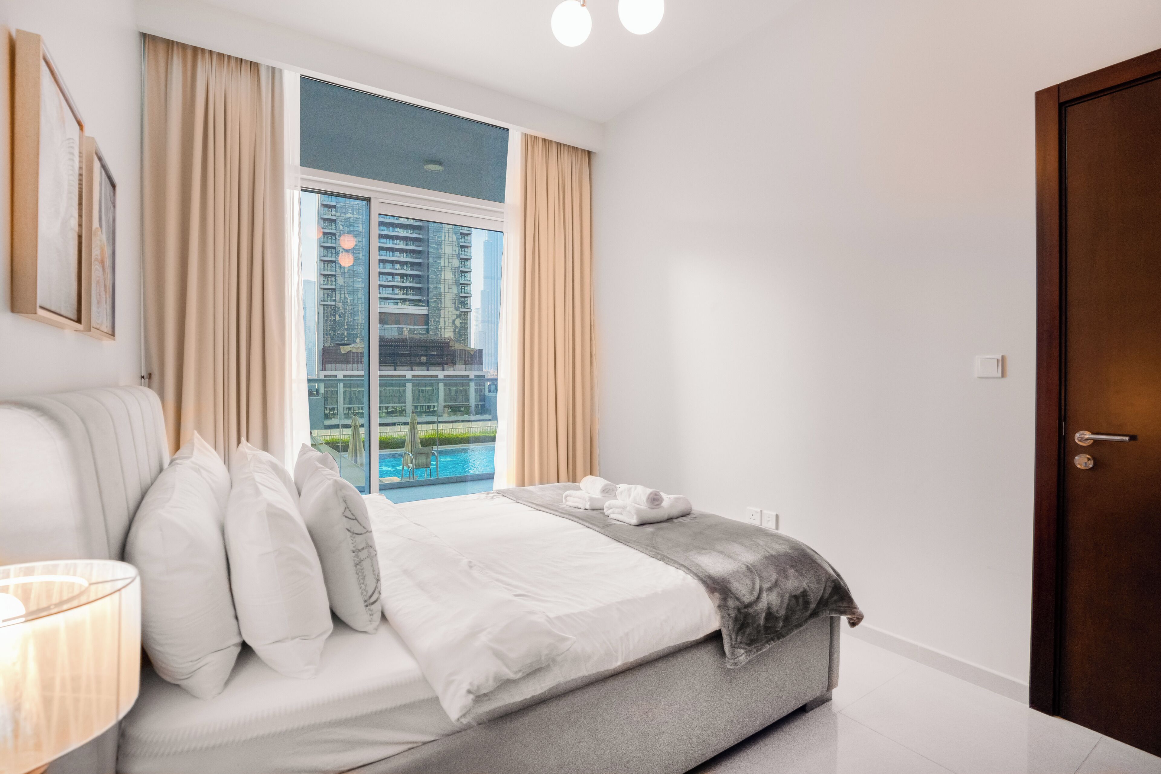 A modern hotel room with a large bed, a view of a cityscape, and a door to the right.