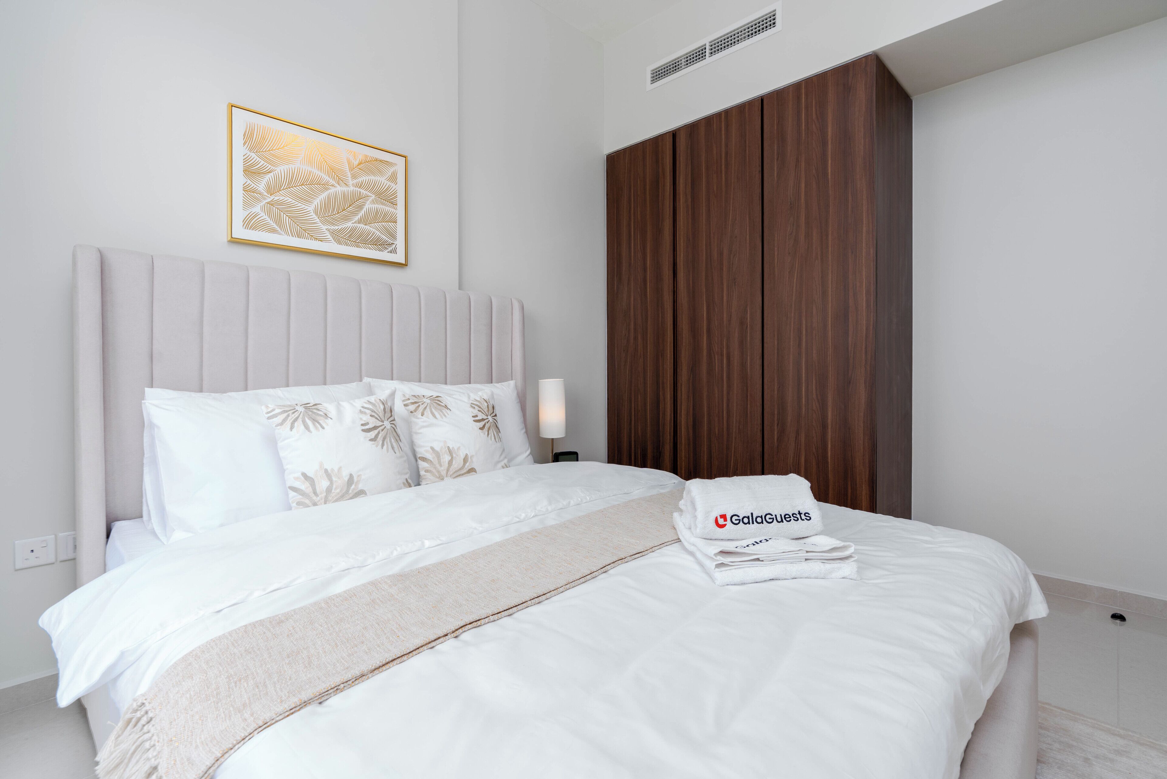 Une chambre d’hôtel comprenant un lit, une tête de lit, une œuvre d’art accrochée au mur, une armoire en bois et une serviette pliée ornée d’un logo.
