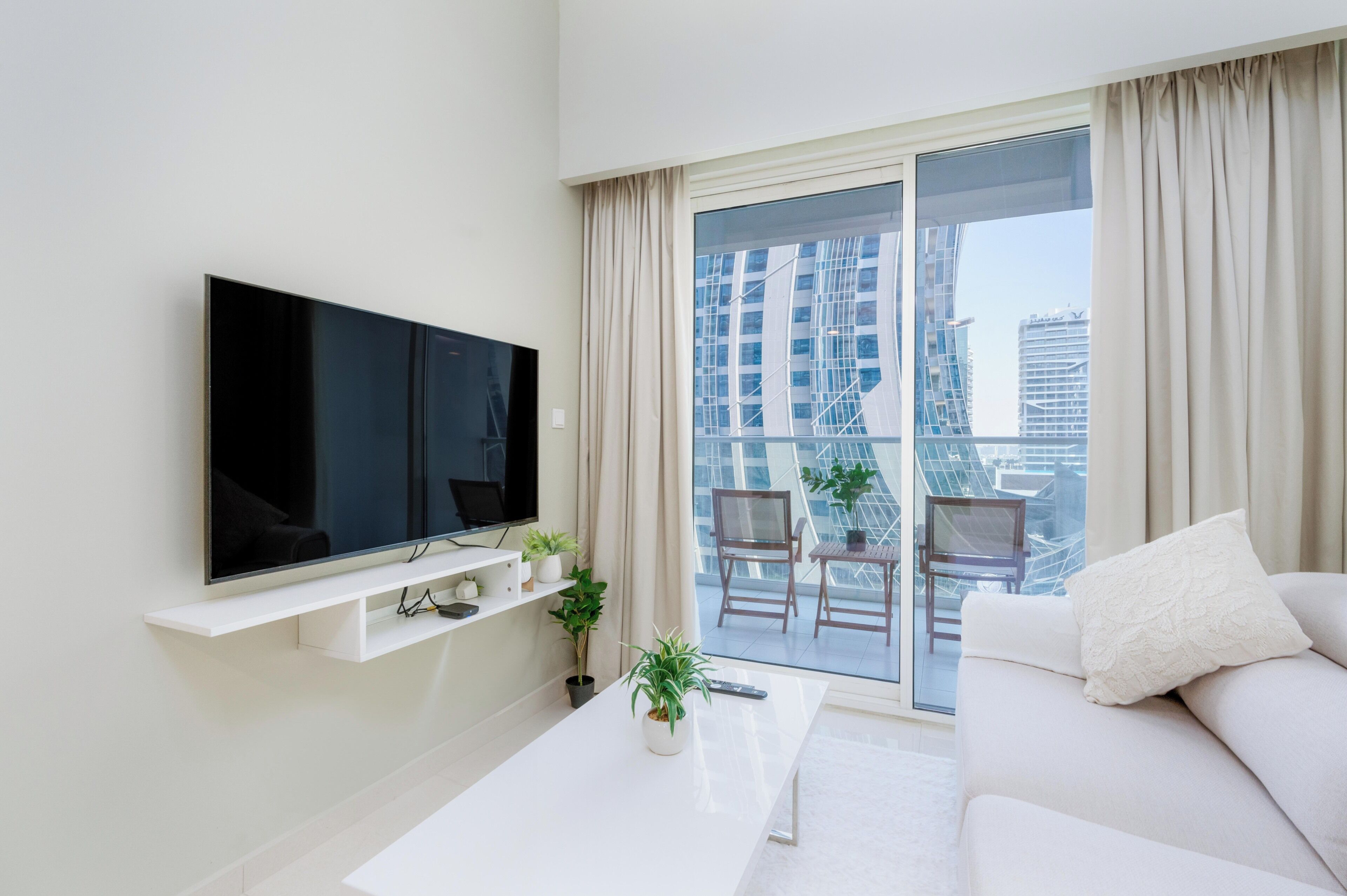 A modern living room with a flat-screen TV, a white coffee table, a sofa, and a balcony with a view of tall buildings.