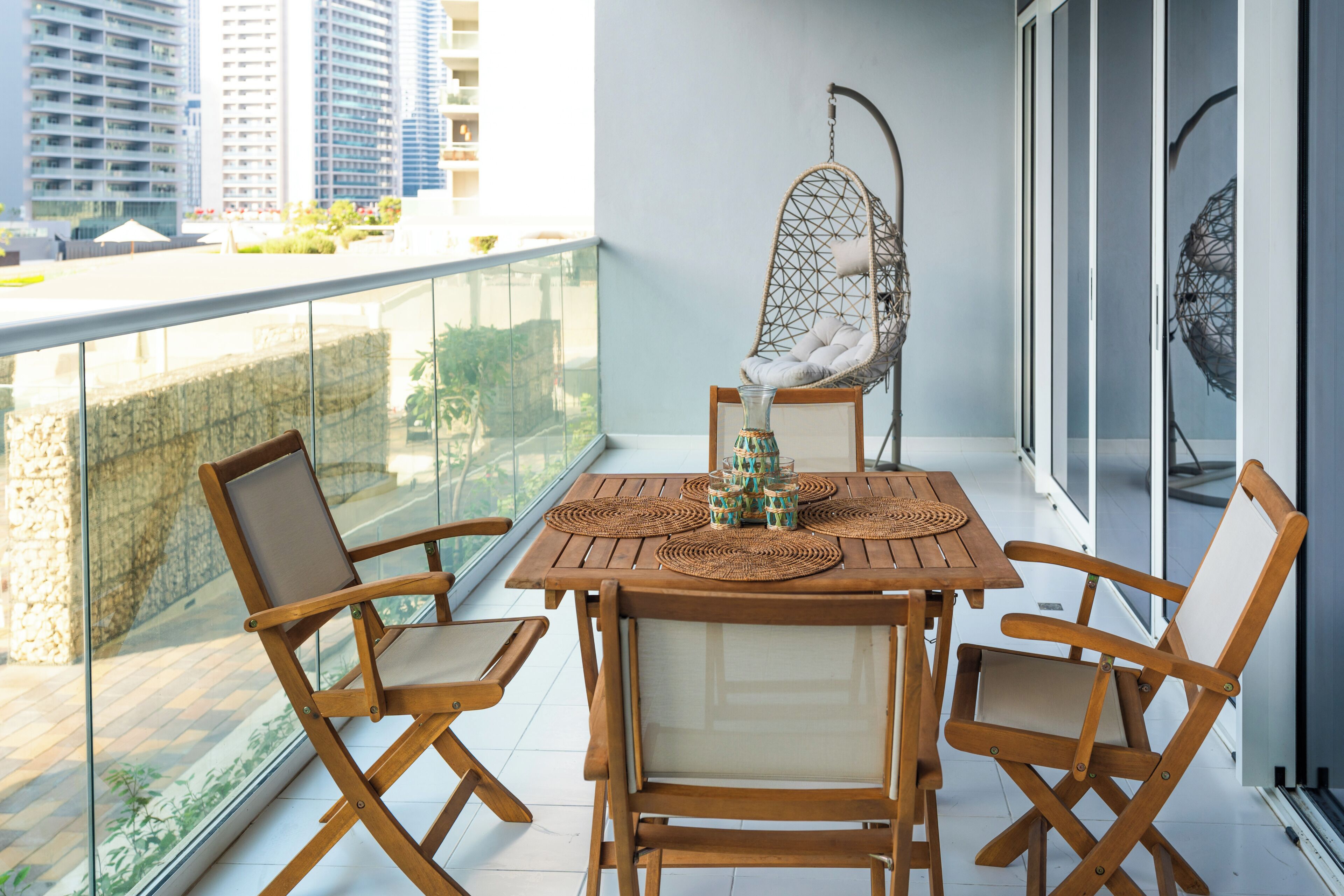 A balcony with a wooden table, four wooden chairs, a hanging chair, and a view of tall buildings.