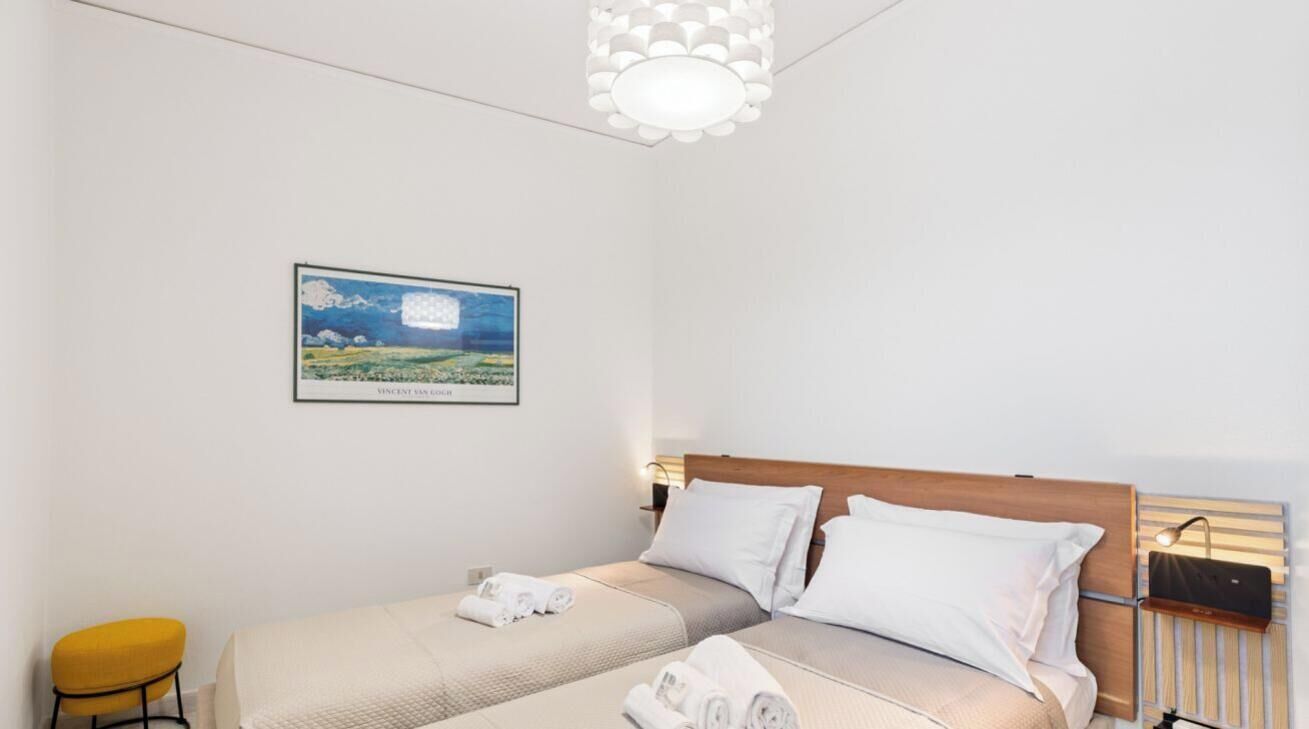 Habitación de hotel con dos camas, un cuadro enmarcado en la pared, una silla amarilla y una mesita de noche con una lámpara.