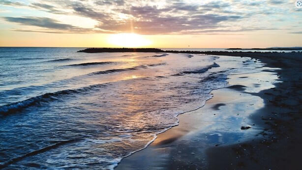 Een strand met golven, een zonsondergang en een landmassa in de verte.
