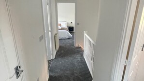 Ein schmaler Flur mit Teppichböden, weißen Wänden und einer Tür zu einem Schlafzimmer mit Bett und Nachttisch.