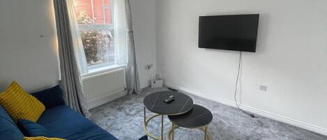 Ein Wohnzimmer mit einer Couch, zwei Beistelltischen, einem an der Wand montierten Fernseher und einem Fenster mit Vorhängen.