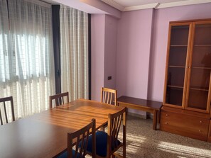 Un comedor con una mesa y sillas de madera, un banco, un mueble con puertas de cristal y cortinas ligeras en las ventanas.