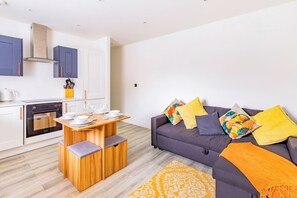 A modern living room with a sofa, a dining table with chairs, a kitchen area with cabinets and an oven, and a patterned rug. - Modern City Centre Flat (Norwich)