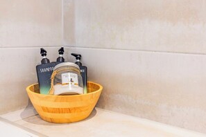 A wooden bowl containing a jar of shampoo and two bottles of body wash on a bathroom shelf. - Modern City Centre Flat (Norwich)
