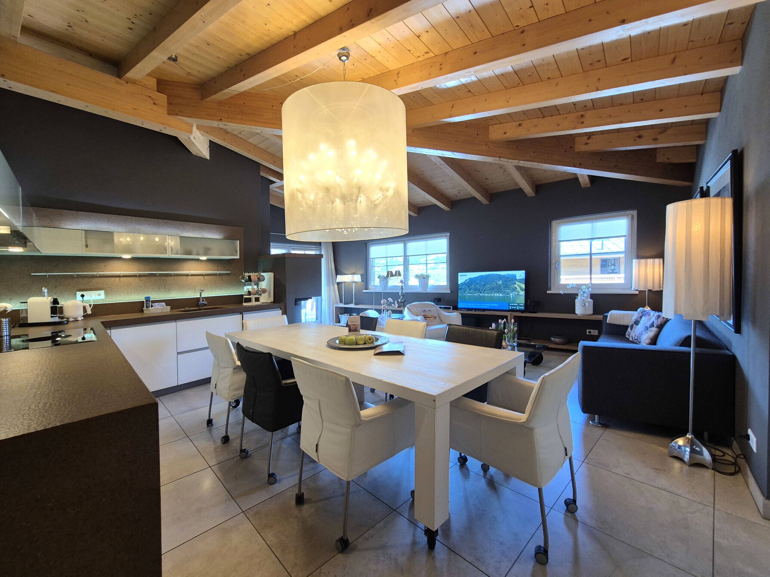 Eine moderne Küche mit Kochinsel, ein Essbereich mit weißem Tisch und Stühlen, eine Wohnbereich mit Sofa und Fernseher sowie ein großes Fenster für natürliches Licht.