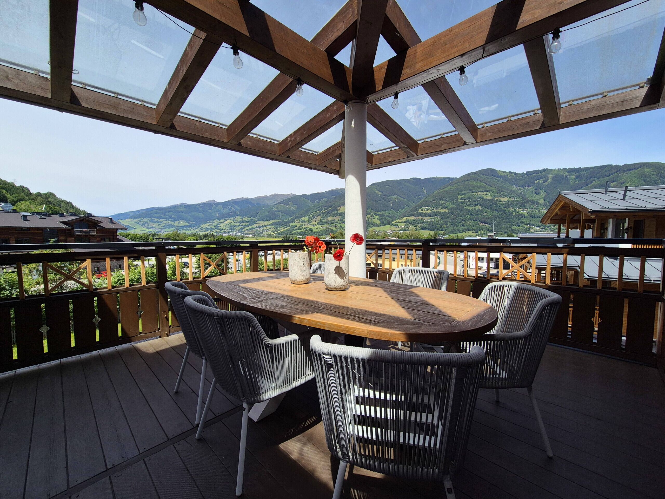 Eine Holzterrasse mit rundem Tisch und Stühlen, einer Glasdecke und Blick auf Berge.