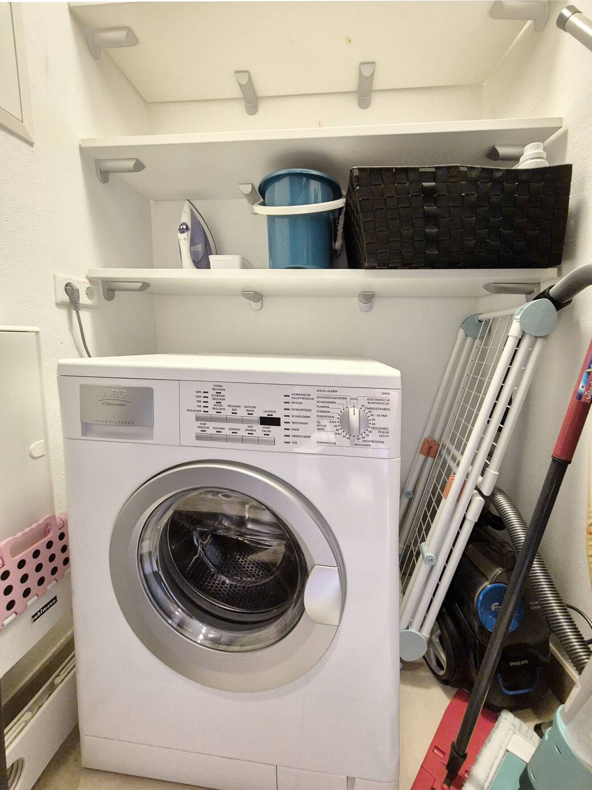 Ein kleiner Hauswirtschaftsraum mit einer Waschmaschine, einem Regal mit einem blauen Behälter und einem Mopp.