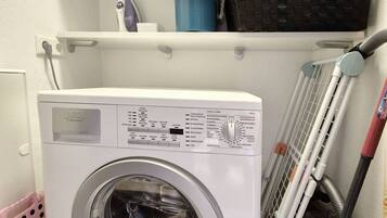 A compact laundry room with a front-loading washing machine, a shelf with a blue container, and a mop.