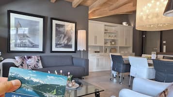 Un salon moderne avec un canapé, une table basse en verre et un coin repas comprenant une table et des chaises blanches. Des tableaux encadrés ornent le mur, et une lampe est posée sur une table d’appoint.