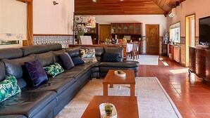 Een ruime woonkamer met een houten plafond, een hoekbank, een salontafel en een keuken op de achtergrond.