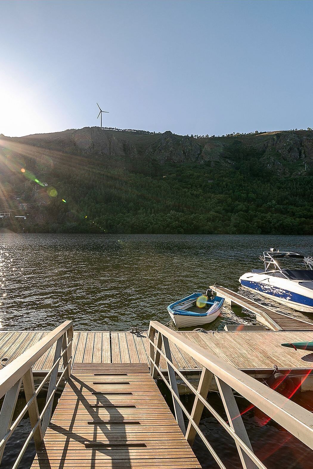 Eine Holzstege, der sich in ruhiges Wasser erstreckt, mit anlegenden Booten, einem klaren Himmel und einer Windkraftanlage im Hintergrund.
