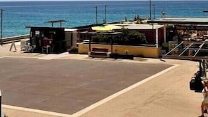 Rannikonäkymä, jossa taivas on kirkas, lipussa liehuu mastossa ja rakennuksessa on parveke, jolta avautuu näkymä merelle.