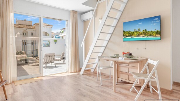 A modern room with a sliding glass door leading to an outdoor area, a white desk with chairs, a ladder, and a beach-themed painting on the wall. - Charming 1BR in Pueblo Mijitas with Terrace by Mijas Golf (Mijas)