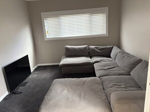 Ein Zimmer mit Ecksofa, Flachbildfernseher und einem Fenster mit Jalousien.