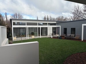 Ein modernes, zweistöckiges Haus mit großem Glasflureingang, einer Terrasse mit Gartenmöbeln und einem gepflegten Rasen.
