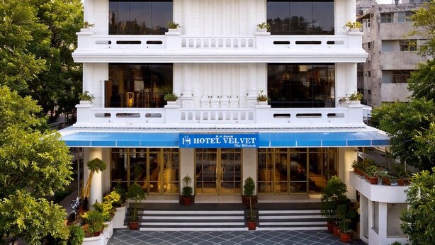 A multi-story hotel with a white facade, a blue awning, and a central entrance with steps. - Hotel Velvet The Ultimate (Prayagraj)
