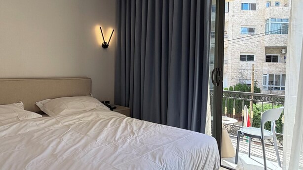 A hotel room with a bed, a nightstand, a wall-mounted lamp, a window with curtains, and a view of the outside building. - 210 Tara-jre Jabal Al Lweibdeh With Stunning View (Amman)
