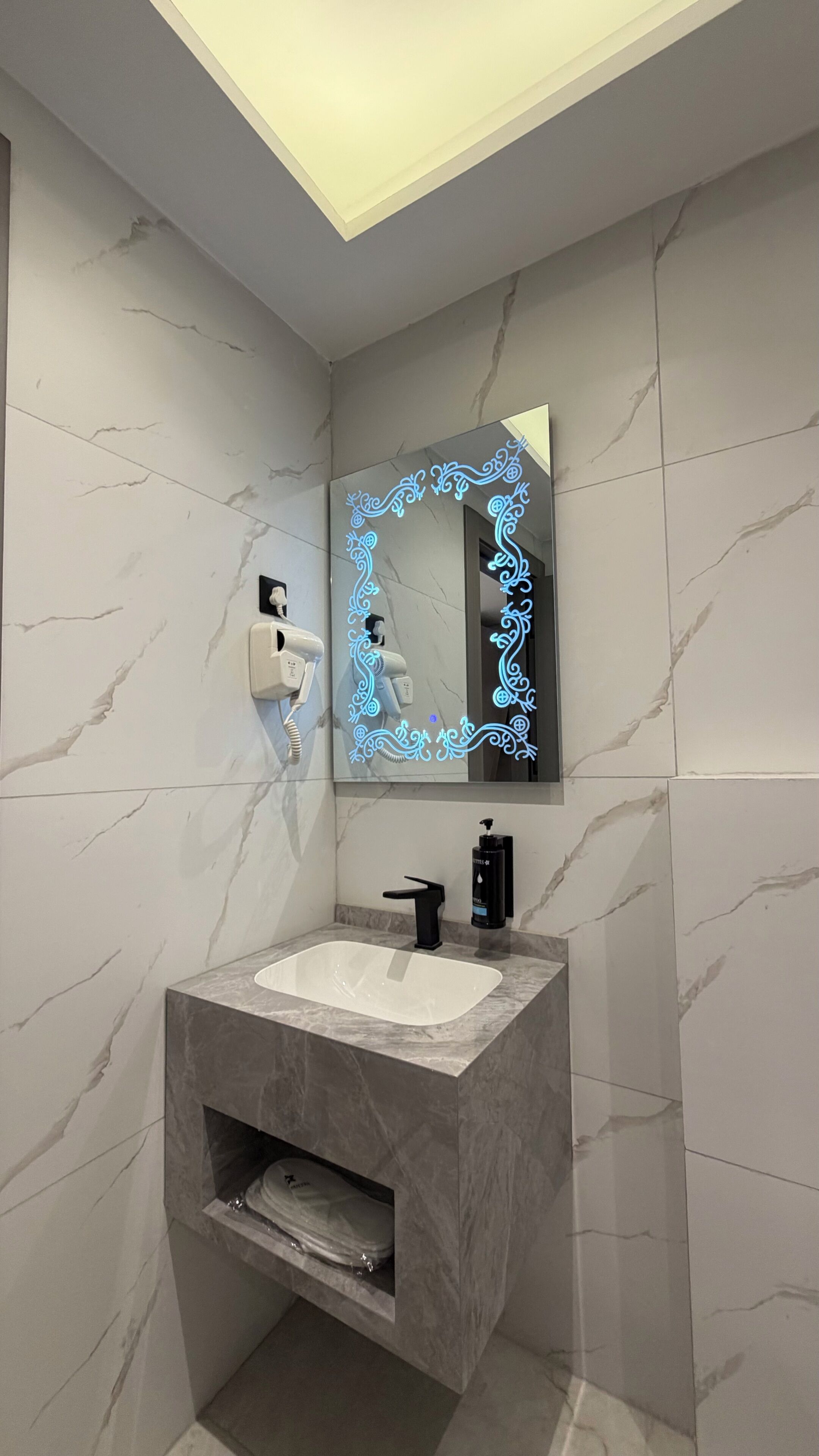 Une salle de bain dotée d’un meuble-lavabo en marbre, d’un miroir mural agrémenté d’un éclairage décoratif, d’un sèche-cheveux et d’un sèche-mains.