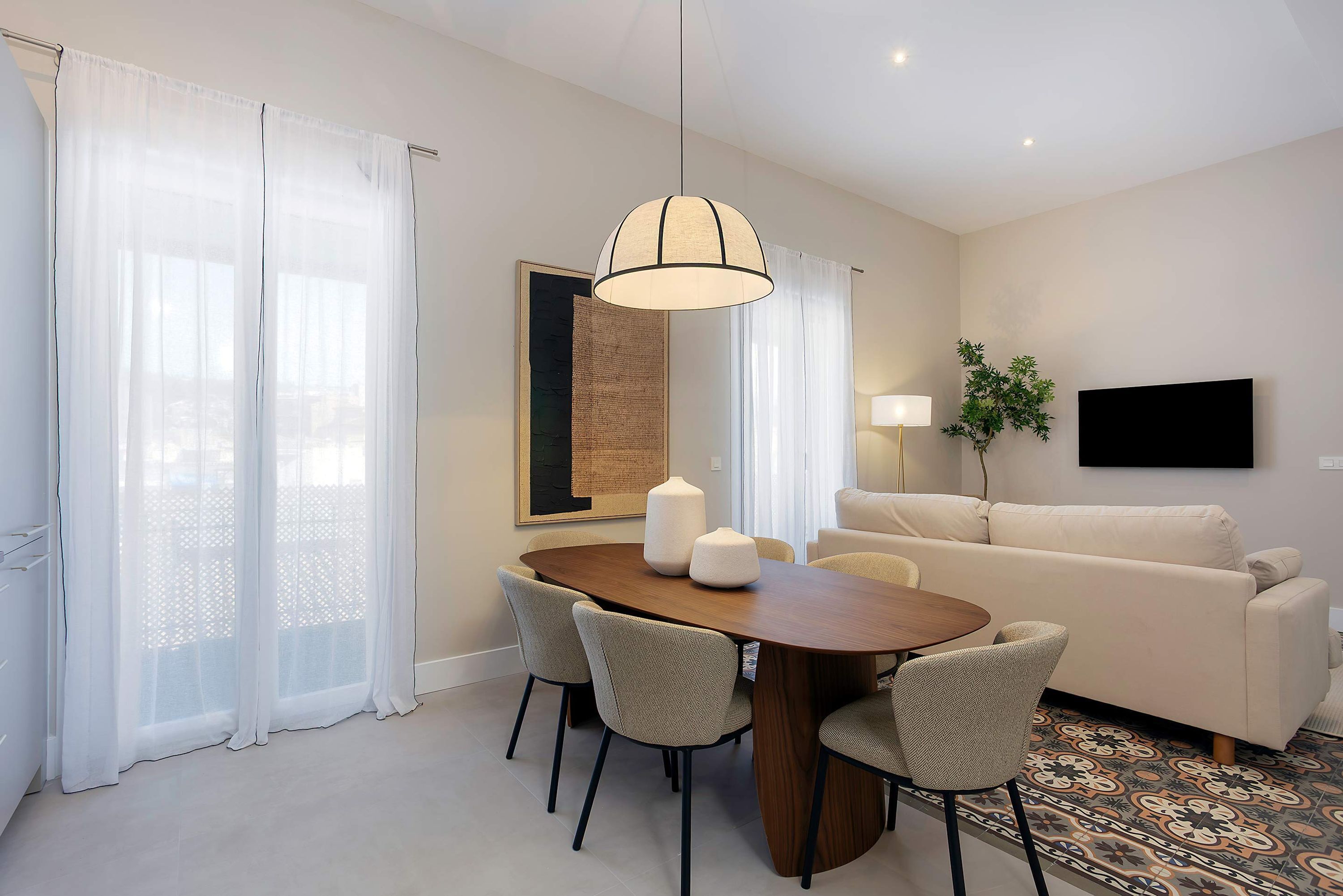 A modern living room with a wooden dining table, four chairs, a sofa, a TV, and a large window with sheer curtains.