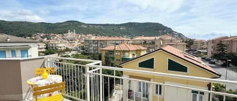A balcony with a yellow chair and table, overlooking a town with buildings and a hill in the background.