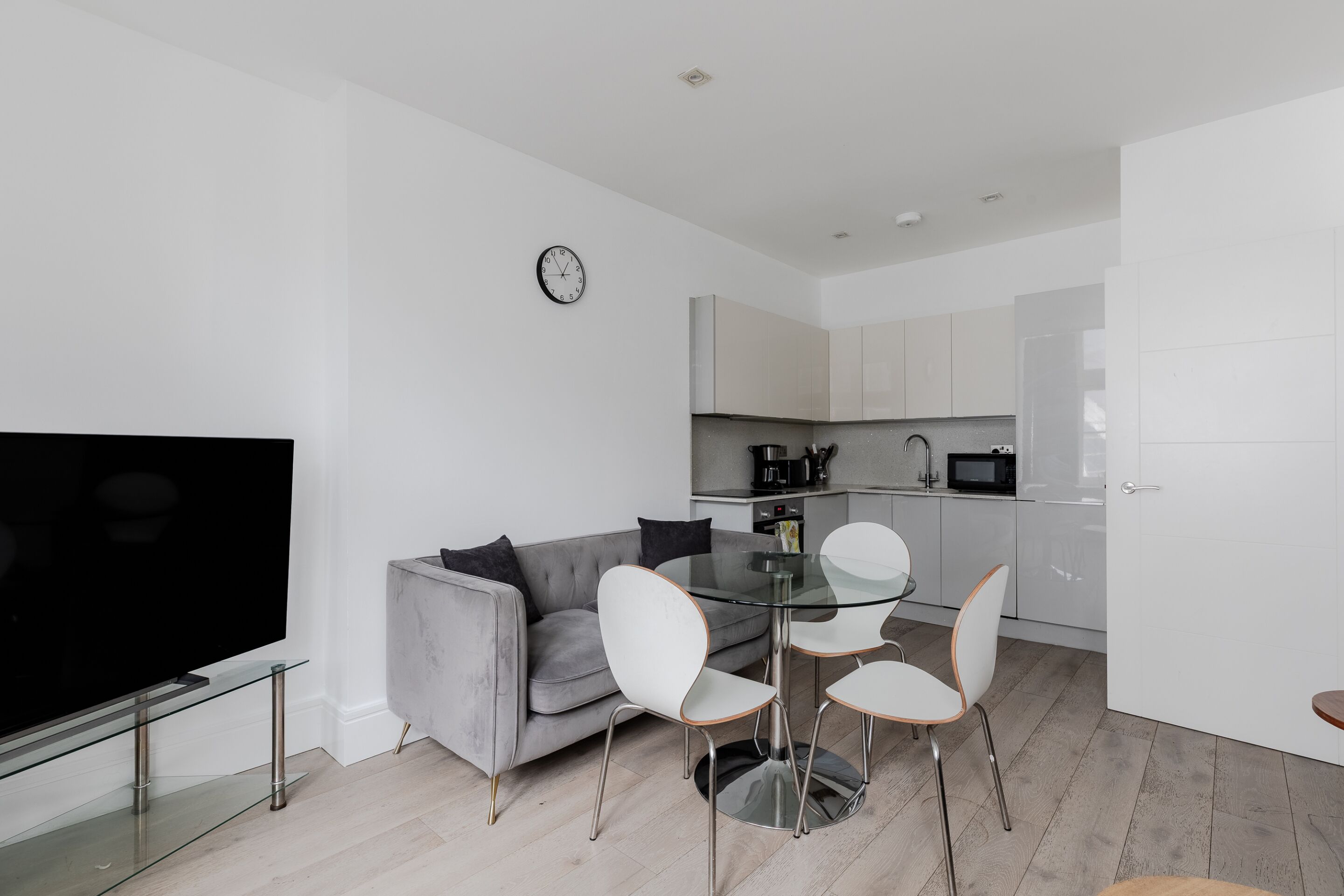 Ein moderner Wohnraum mit Flachbildfernseher, grauem Sofa, Glastisch mit weißen Stühlen und Küchenzeile mit weißen Schränken und Geräten.