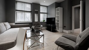 A modern living room with a sectional sofa, a glass coffee table, a grey armchair, and a flat-screen TV. - Superb 2 Bedroom Apartments (London)