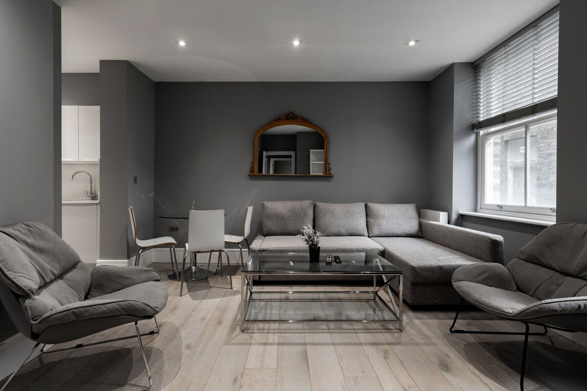 A modern living room with a grey sofa, two armchairs, a glass coffee table, and a mirror on the wall.