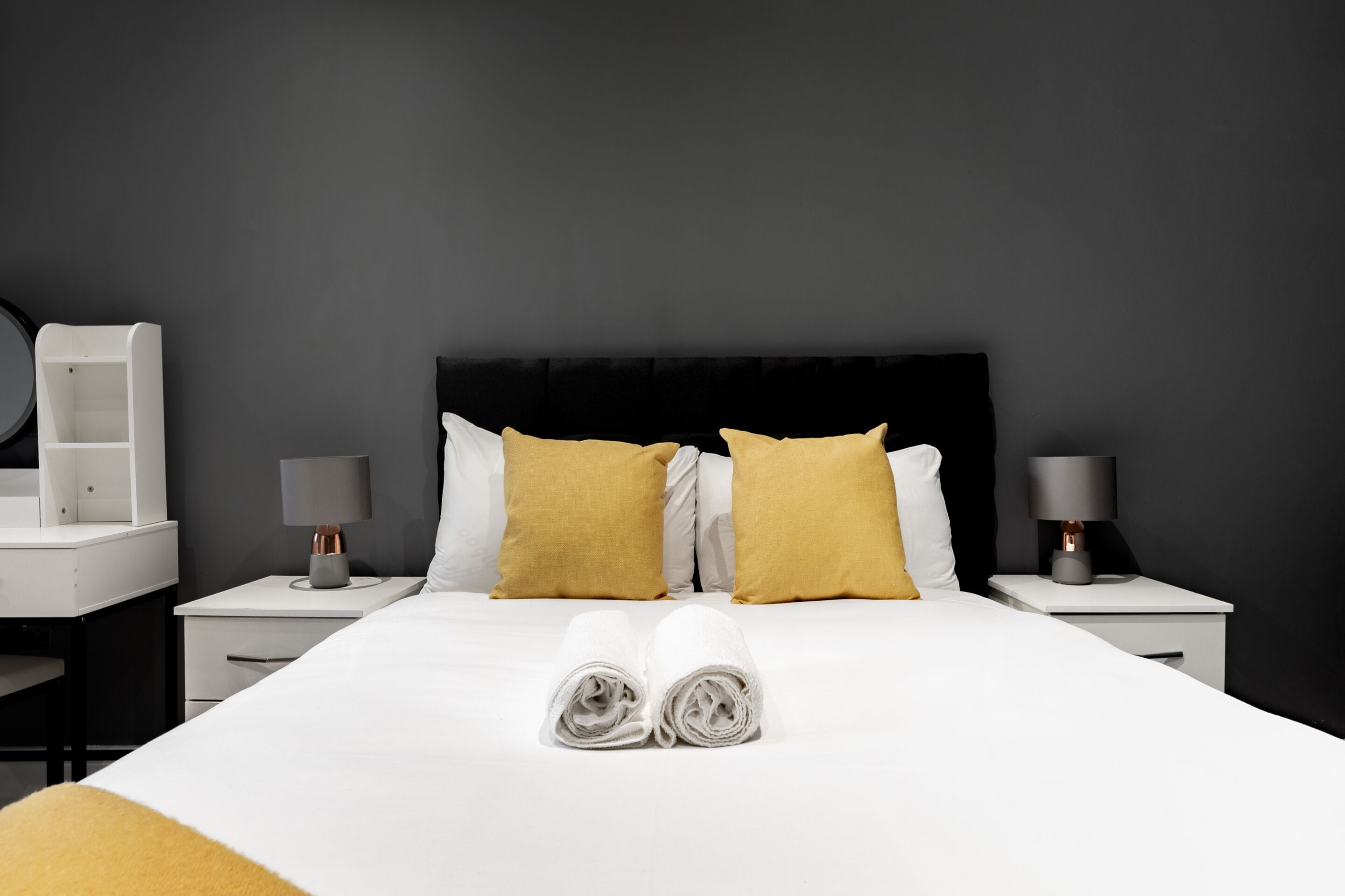 A neatly made bed with two pillows, a bedside table with a lamp, and a nightstand with a lamp.