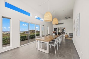 Un comedor amplio con una larga mesa de madera, sillas blancas y ventanas grandes que ofrecen vistas al exterior.
