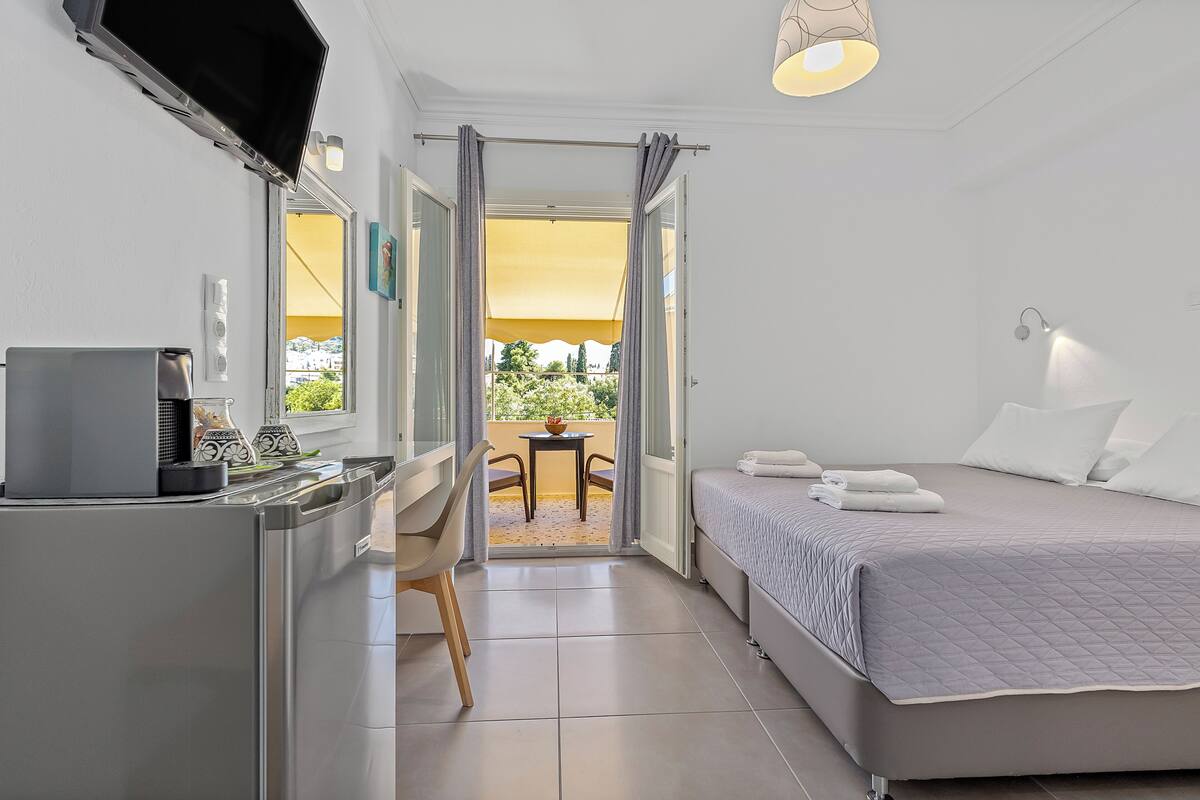 Una habitación de hotel con una cama, un escritorio con teléfono, una silla, una mesa con planta y una puerta abierta que da a un área exterior con mesa y sillas.