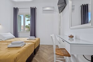 A hotel room with two beds, a desk, a chair, an air conditioning unit, and a window with curtains. - Kamelia Hotel (Spetses)