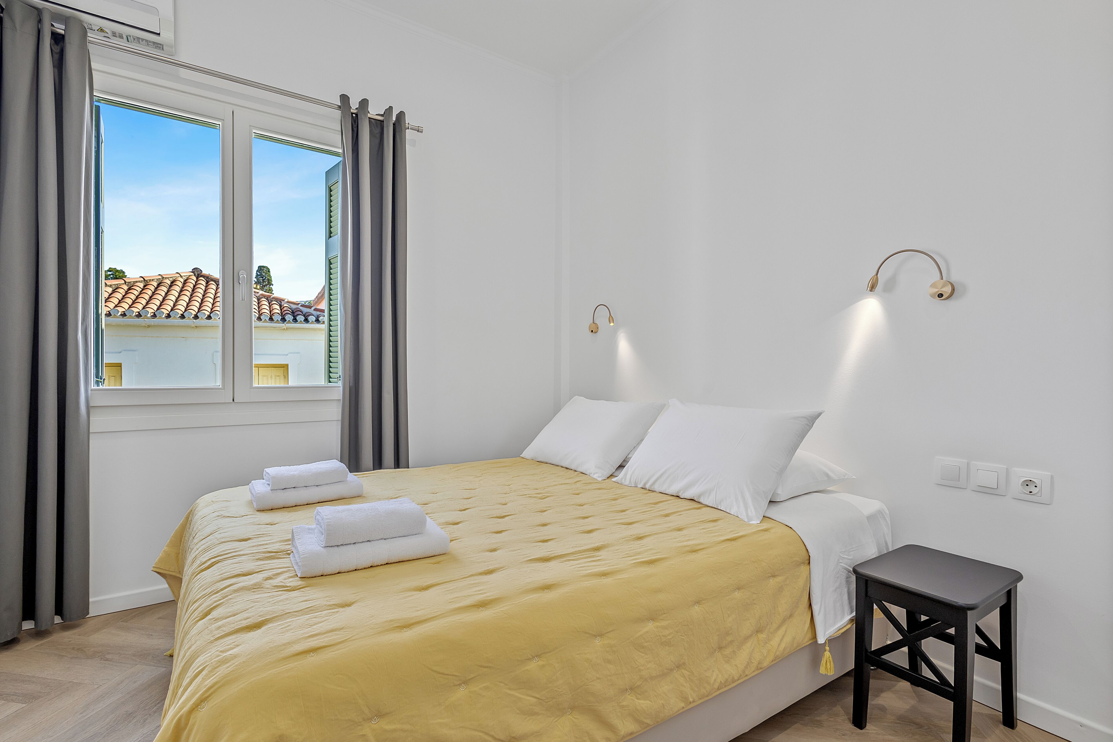 A bedroom with a bed, a small table, a window with curtains, and a wall-mounted light fixture.