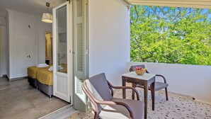 A balcony with a table and chairs, a bed with a yellow bedspread, and a view of green trees.