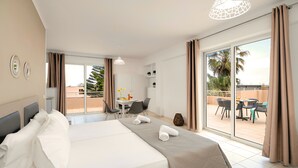 Hotellihuone, jossa on sänky, ruokapöytä tuoleineen, parveke pöytineen ja tuoleineen sekä näkymä palmuille.