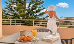 A balcony with a table set for breakfast, including croissants, a coffee cup, and a glass of orange juice. A book titled 'RELAXATION' is also visible.